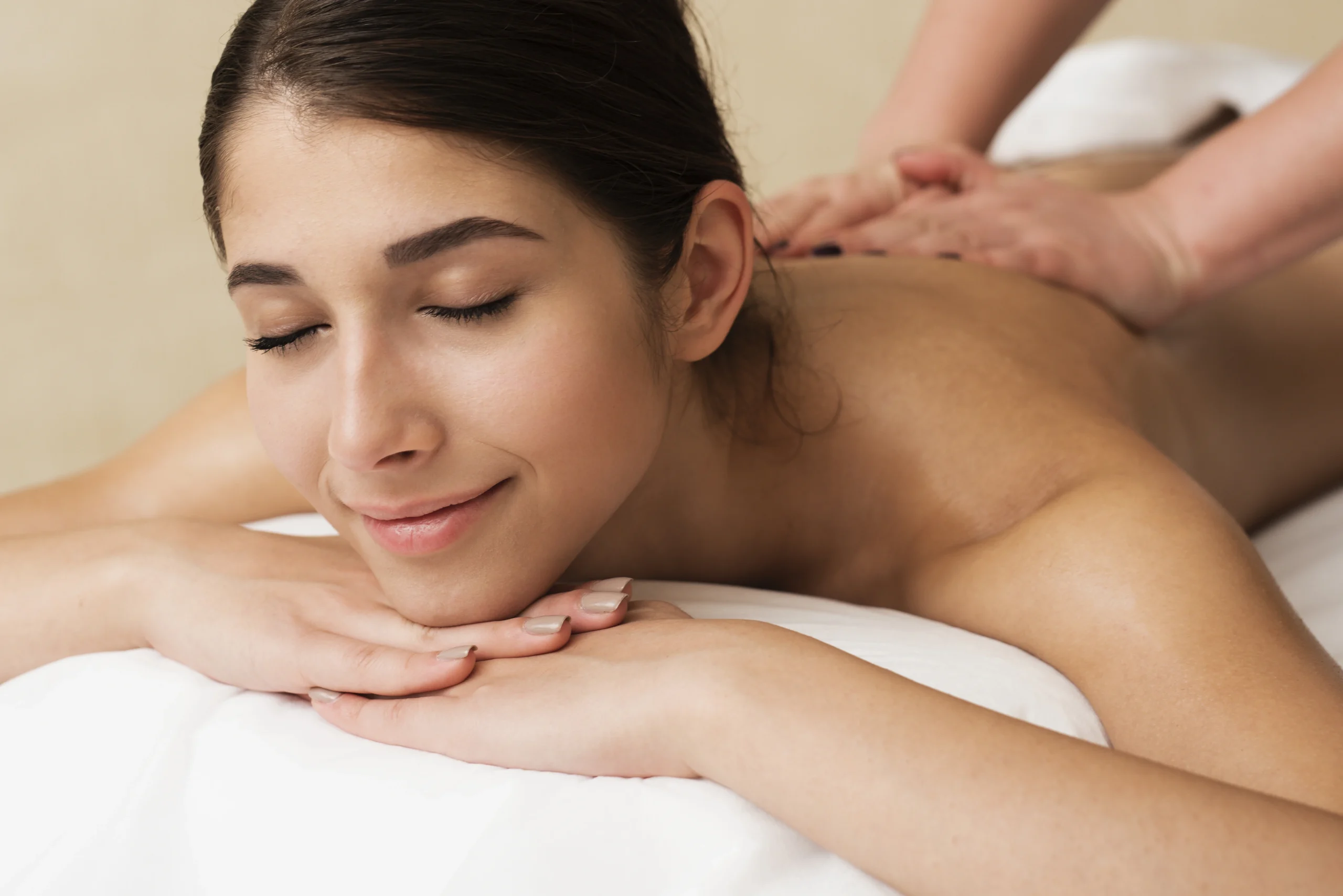 A close-up of a young woman with her eyes closed and a peaceful smile, lying on her stomach while receiving a back massage.