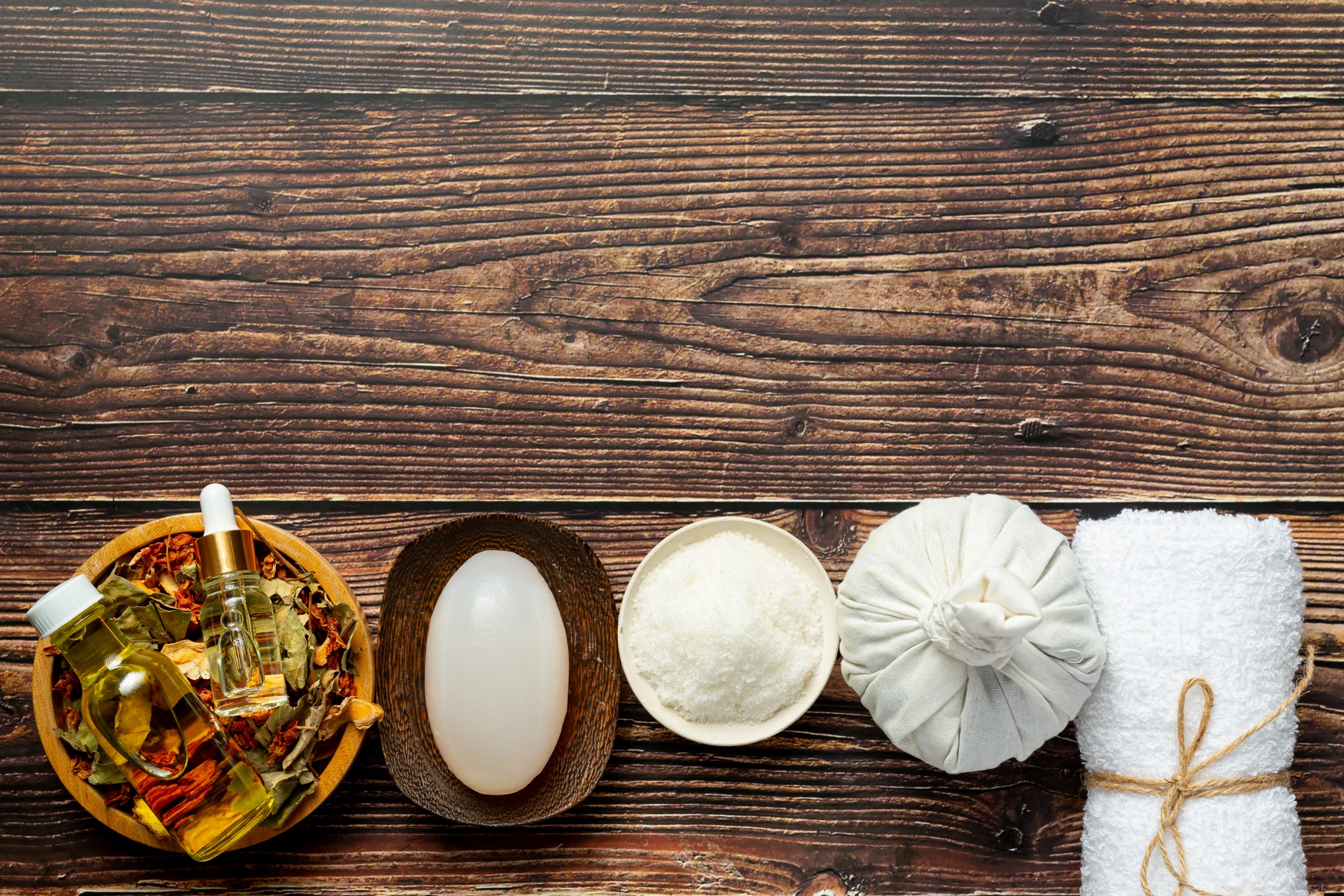 Panchakarma - An overhead shot of various wellness items arranged in a row at the bottom of a dark, rustic wooden surface. The collection includes: A wooden bowl of dried herbs and two essential oil bottles. A smooth, white massage stone in a dark dish. A small bowl of sea salt. A traditional white herbal compress. A rolled white towel tied with twine.