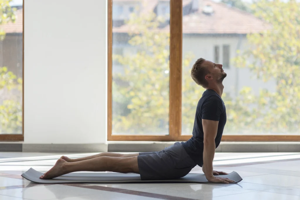Side view of a man in a dark t-shirt and grey shorts performing a cobra stretch on a yoga mat.