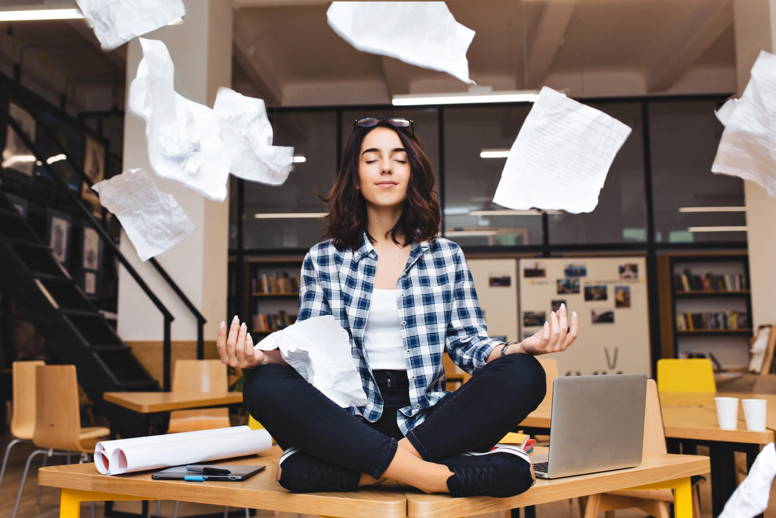 Corporate Wellness- A woman in a plaid shirt sits in a meditative lotus pose on an office desk, eyes closed in calm focus while sheets of white paper fly through the air around her in a modern workspace.