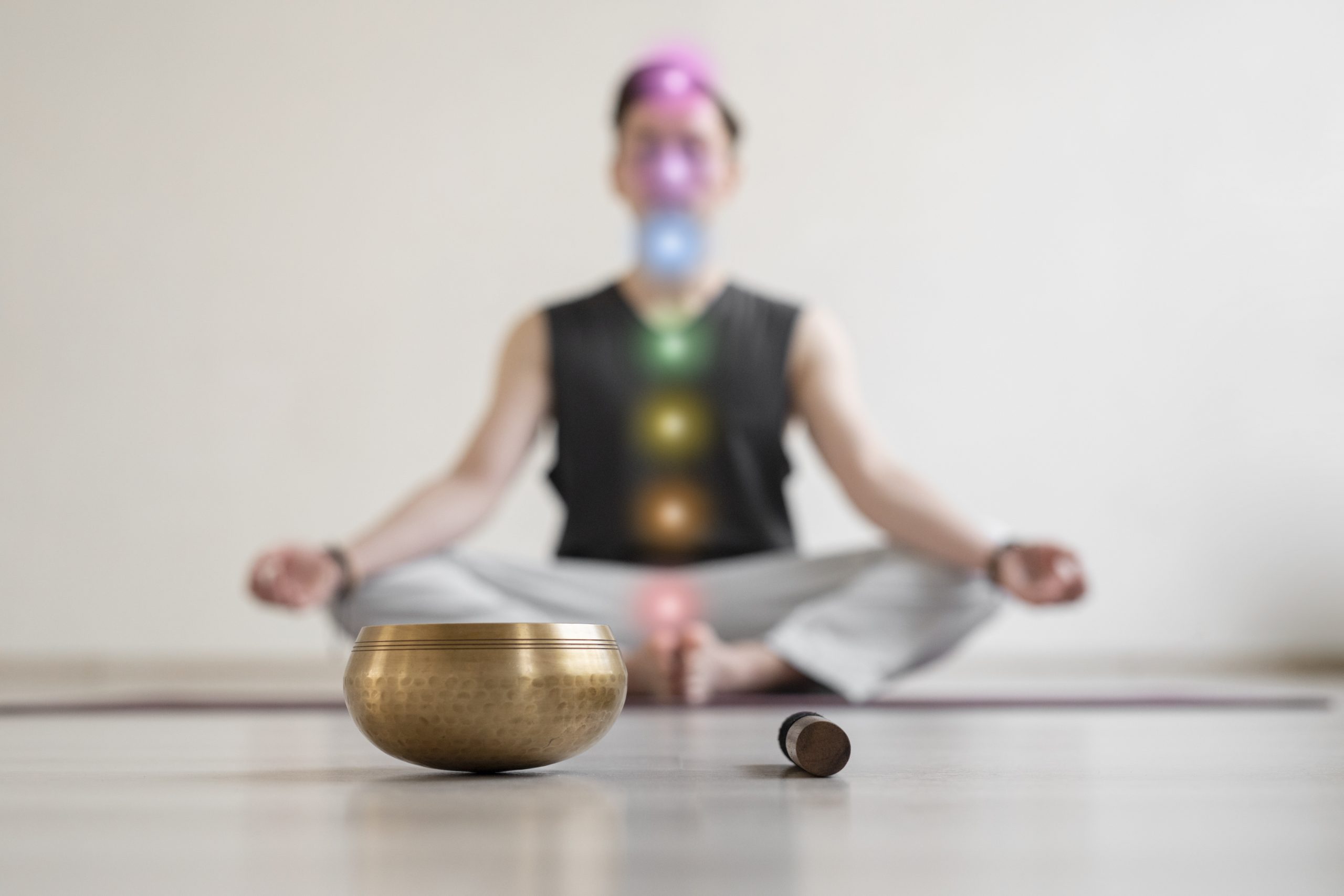Holistic Wellbeing- A blurred, centered shot of a person sitting in a cross-legged meditation pose on a yoga mat, with a sharp-focus brass singing bowl and mallet in the foreground. Seven colorful, glowing circles representing the chakras are overlaid vertically along the person's torso and head, ranging from red at the base to violet at the crown.