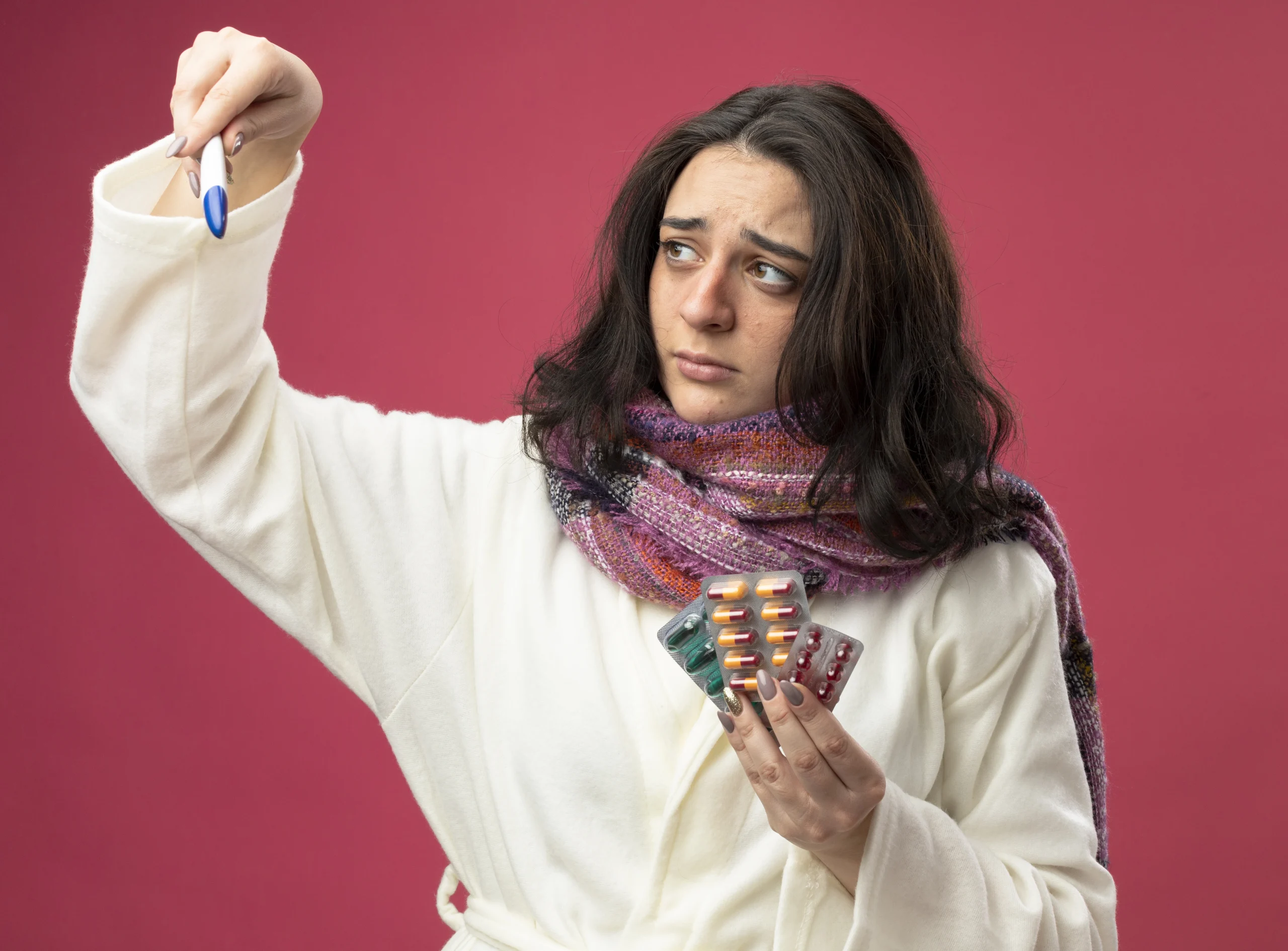 Hormonal Imbalance- A medium shot of a young woman with a concerned expression, wearing a white bathrobe and a thick purple and grey scarf against a solid pink background. She holds up a digital thermometer in her right hand, looking at it with a furrowed brow, while her left hand holds several blister packs of various colored pills and capsules.