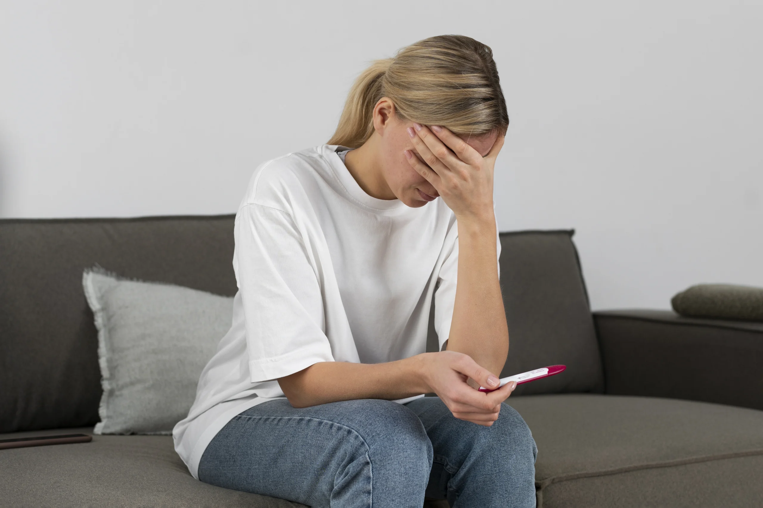 Hormonal Imbalance- A blonde woman sitting on a grey sofa, wearing a white t-shirt and blue jeans. She appears distressed, with her right hand covering her forehead and eyes in a gesture of worry or grief. In her left hand, she holds a pink and white pregnancy test. The background is a simple, neutral-toned living room.
