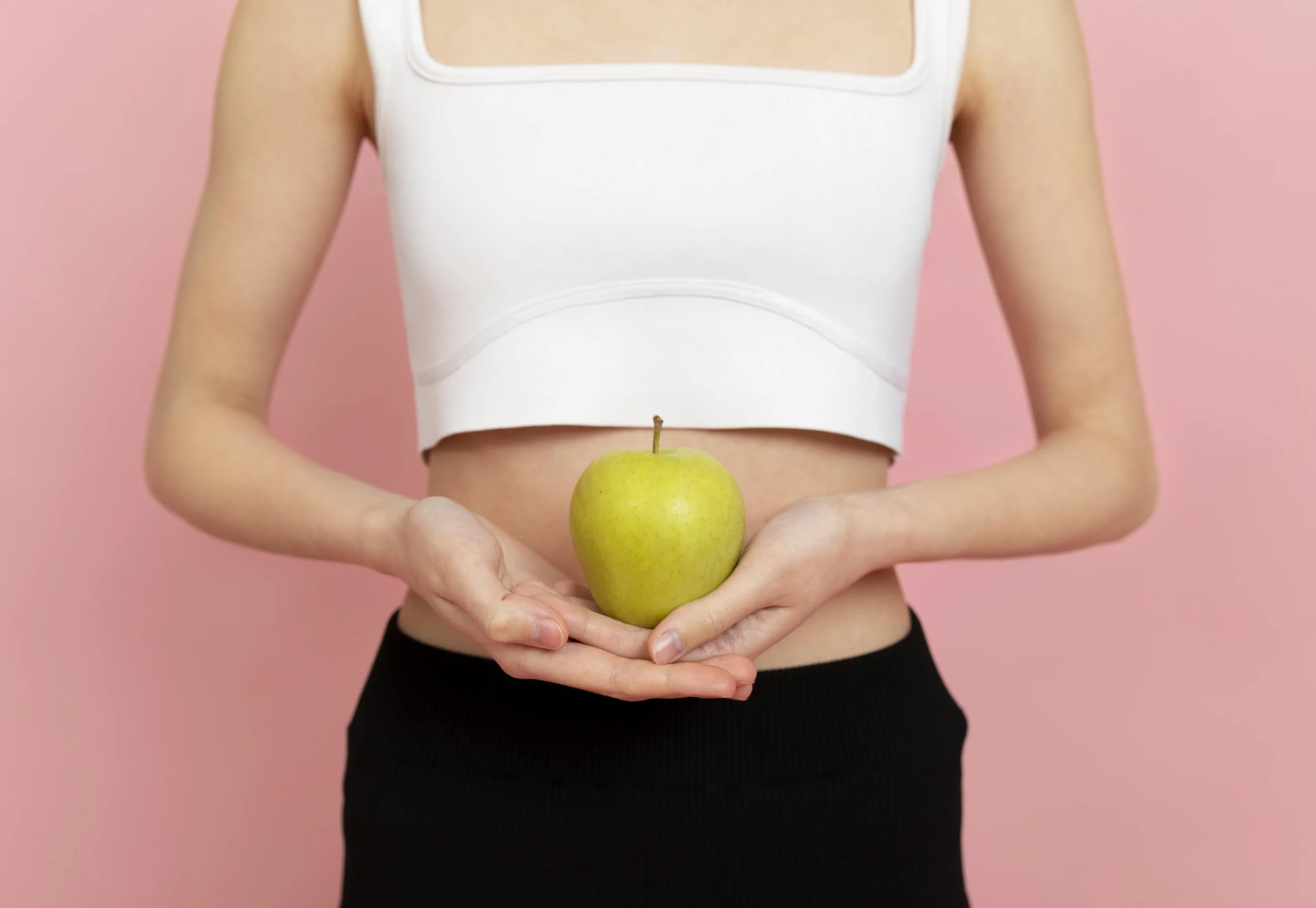 Digestive and Metabolic Health-A medium shot against a solid pink background shows a woman from the chest down to the waist. She wears a white square-neck crop top and black bottoms. She holds a green apple in both hands in front of her midsection.