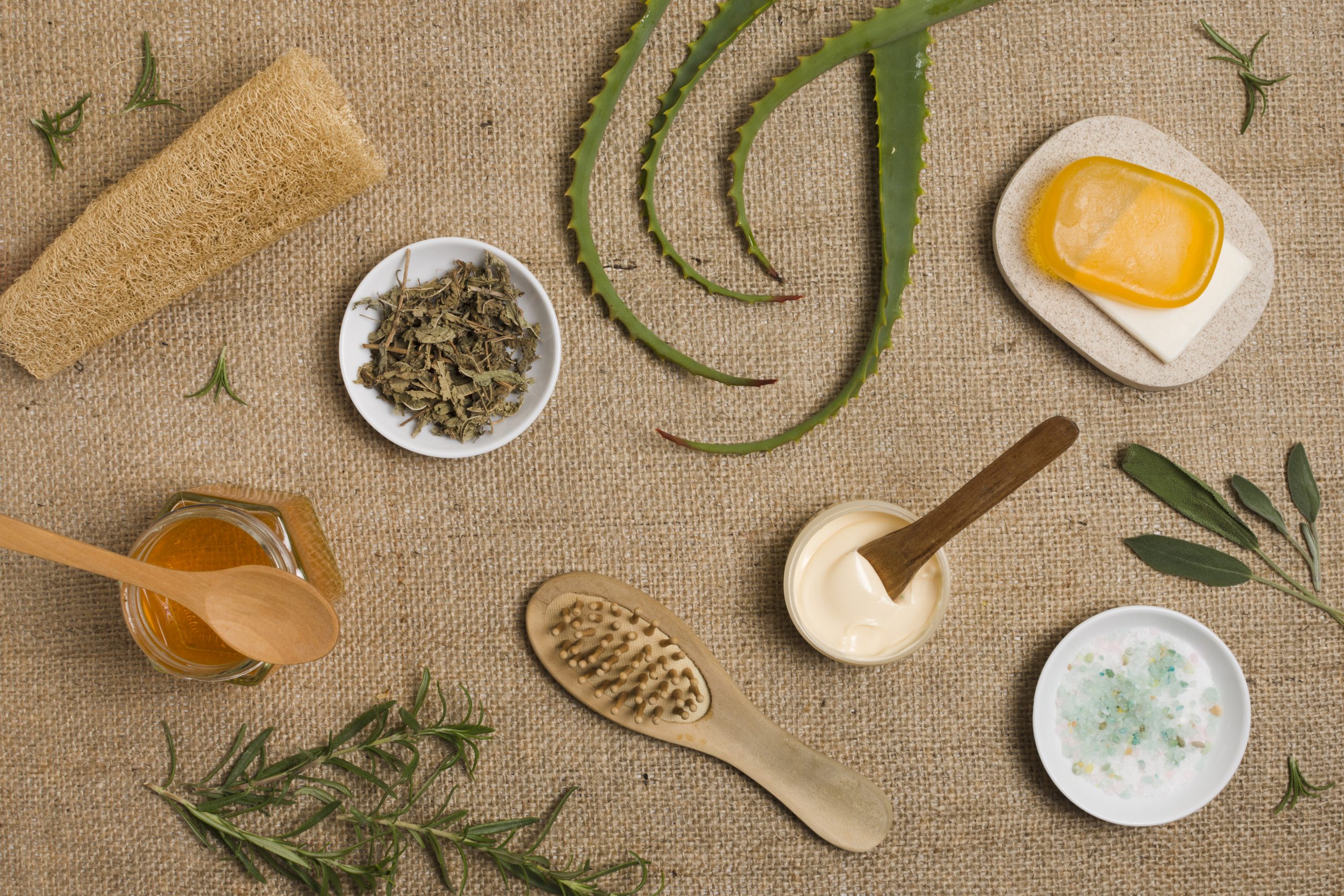 Personal care: An overhead shot shows an assortment of natural skincare and wellness items arranged on a rustic burlap surface. The collection includes: Botanicals: Fresh green aloe vera leaves, sprigs of rosemary, and a small white bowl of dried herbs. Tools: A wooden hairbrush, a loofah sponge, and two small wooden spoons resting in jars. Products: A jar of honey (or golden oil), a small bowl of creamy white salve, a dish of bath salts, and a bar of translucent amber soap on a stone tray. The aesthetic is earthy and organic, emphasizing a holistic or DIY approach to beauty.