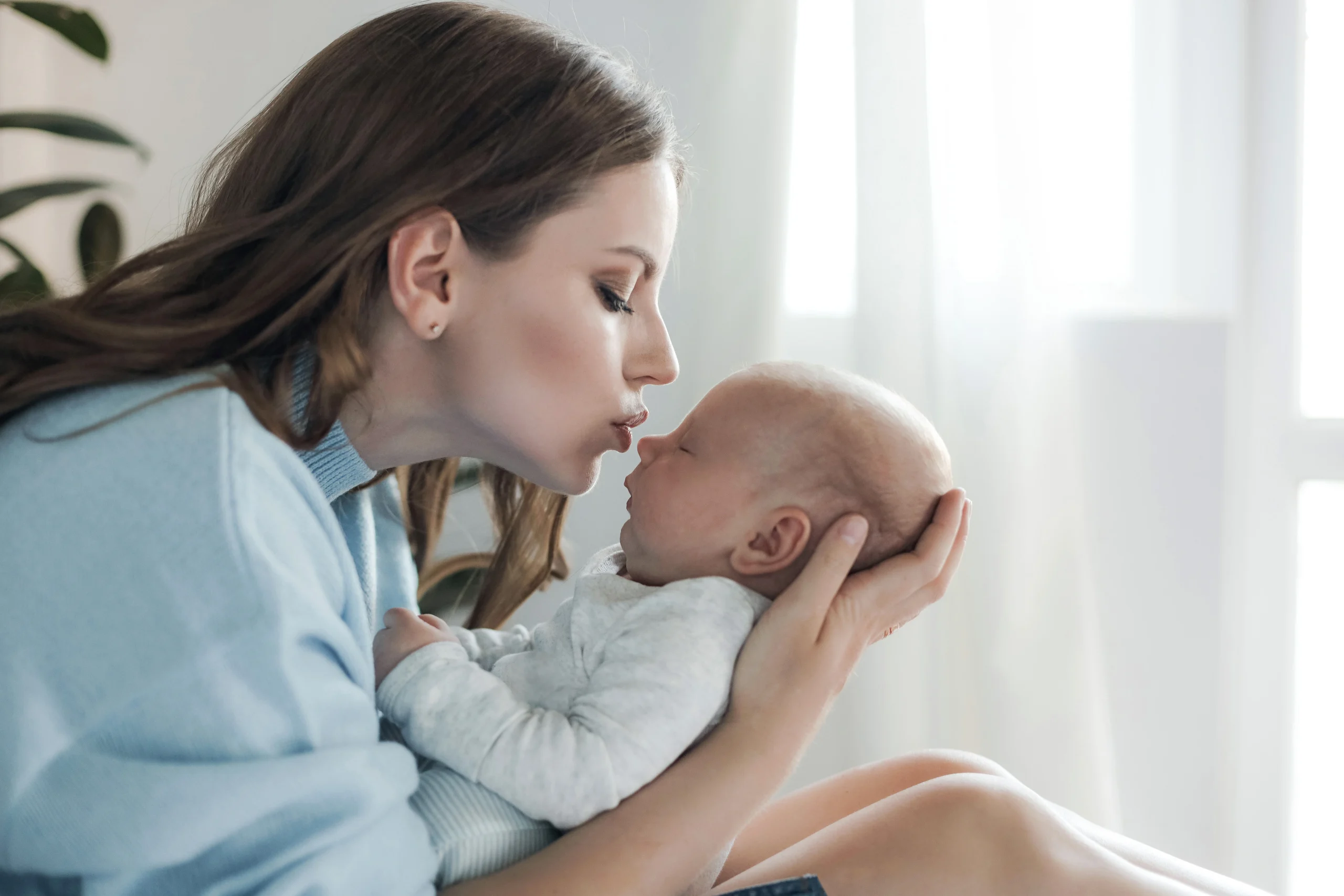 Pre and Postnatal Care- A young woman with long, wavy brown hair, wearing a light blue sweater, leans in to kiss a sleeping newborn baby. She is cradling the infant's head and body gently in her hands. The baby is dressed in a white onesie and appears calm and peaceful. The scene is set in a bright, softly lit room with a blurred window and a green plant in the background, creating a warm and intimate atmosphere.