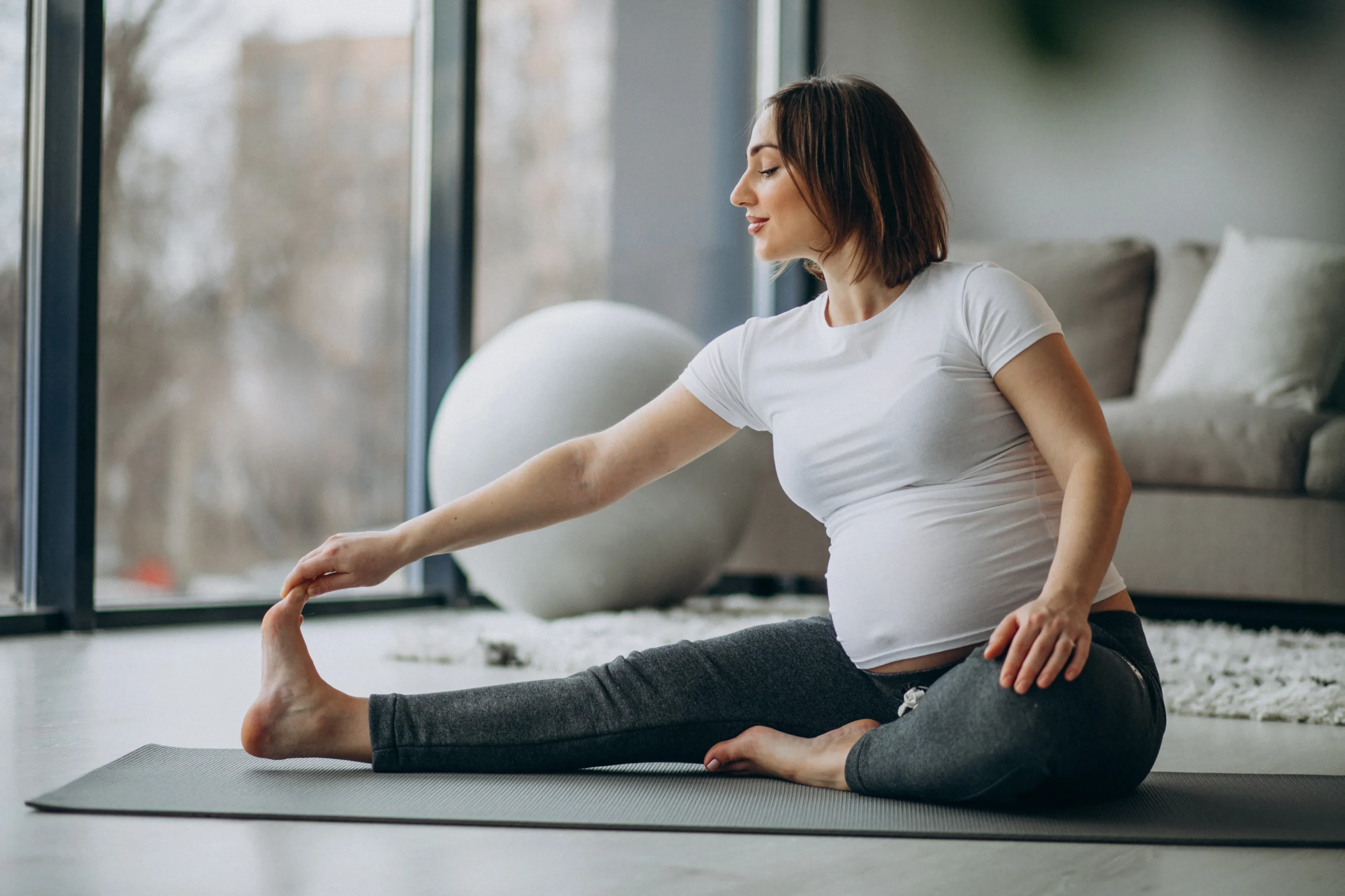 Prenatal and Postnatal Care- A pregnant woman with short brown hair is sitting on a gray yoga mat in a bright, modern living room, performing a seated side stretch. She is wearing a white t-shirt and dark gray leggings. One leg is extended straight out to the side while she reaches for her toes with one hand; her other leg is tucked in toward her body.