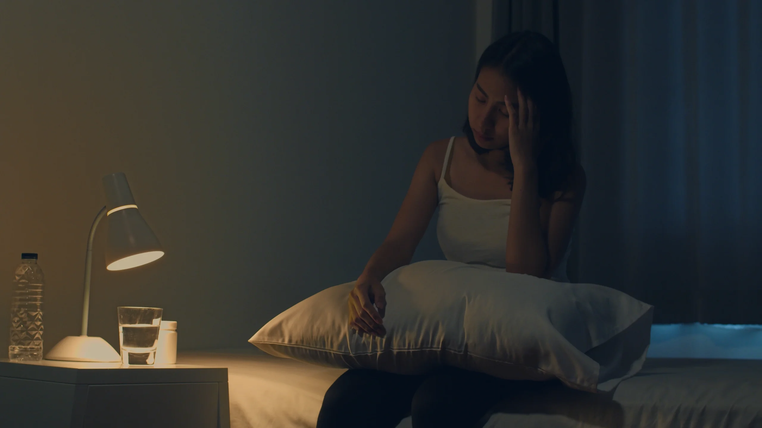 Destress Retreat: A young woman sits on the edge of her bed at night, holding her head in her hand with an expression of distress or exhaustion.