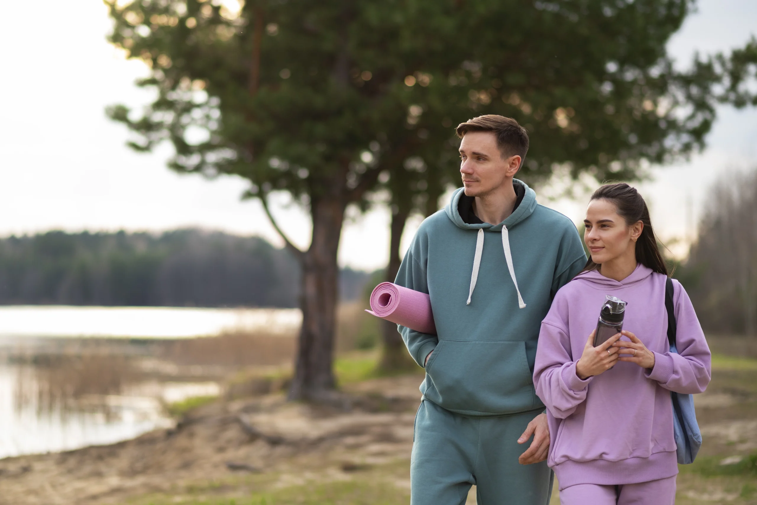 Lifestyle prevention- A young man in a teal tracksuit and a woman in a lavender tracksuit walk along a lakeshore at dusk. He carries a pink yoga mat, and she holds a water bottle. Soft evening light and trees are in the background.