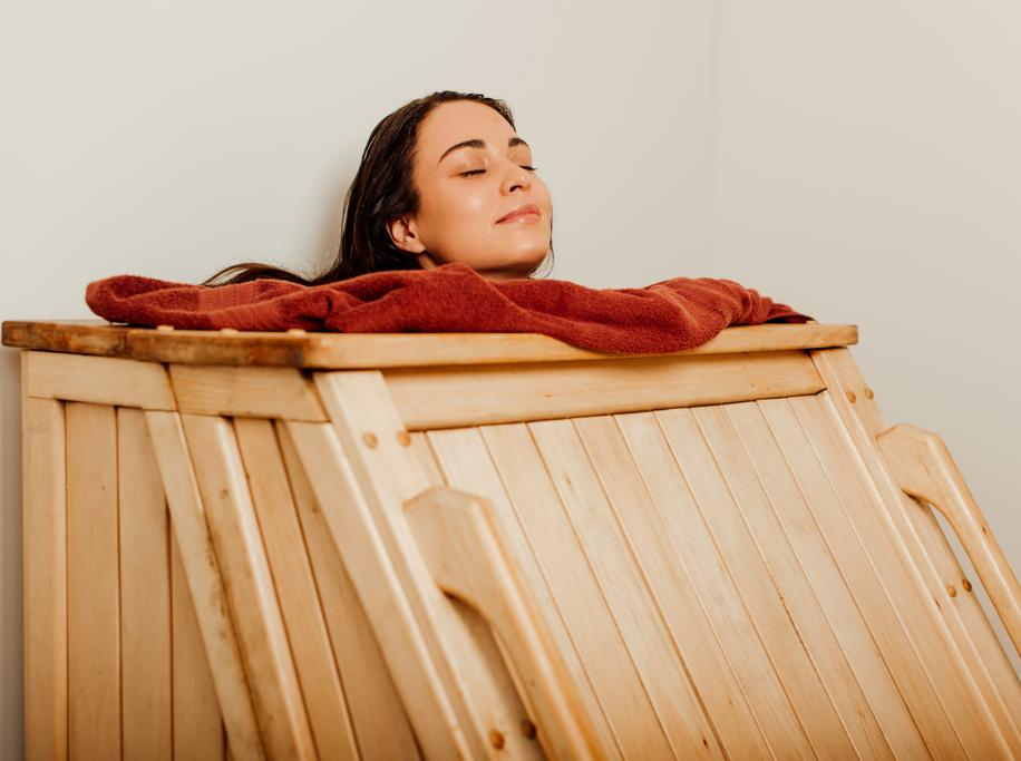 Swedhanam - An eye-level medium shot shows a woman with dark hair and a serene expression resting inside a light-colored wooden steam cabinet. Her eyes are closed and her head is tilted back slightly, resting against a plush, reddish-brown towel draped around the neck opening of the box. The cabinet is constructed from vertical wooden slats, and the warm, soft lighting creates a tranquil atmosphere against the neutral, off-white walls of the room.