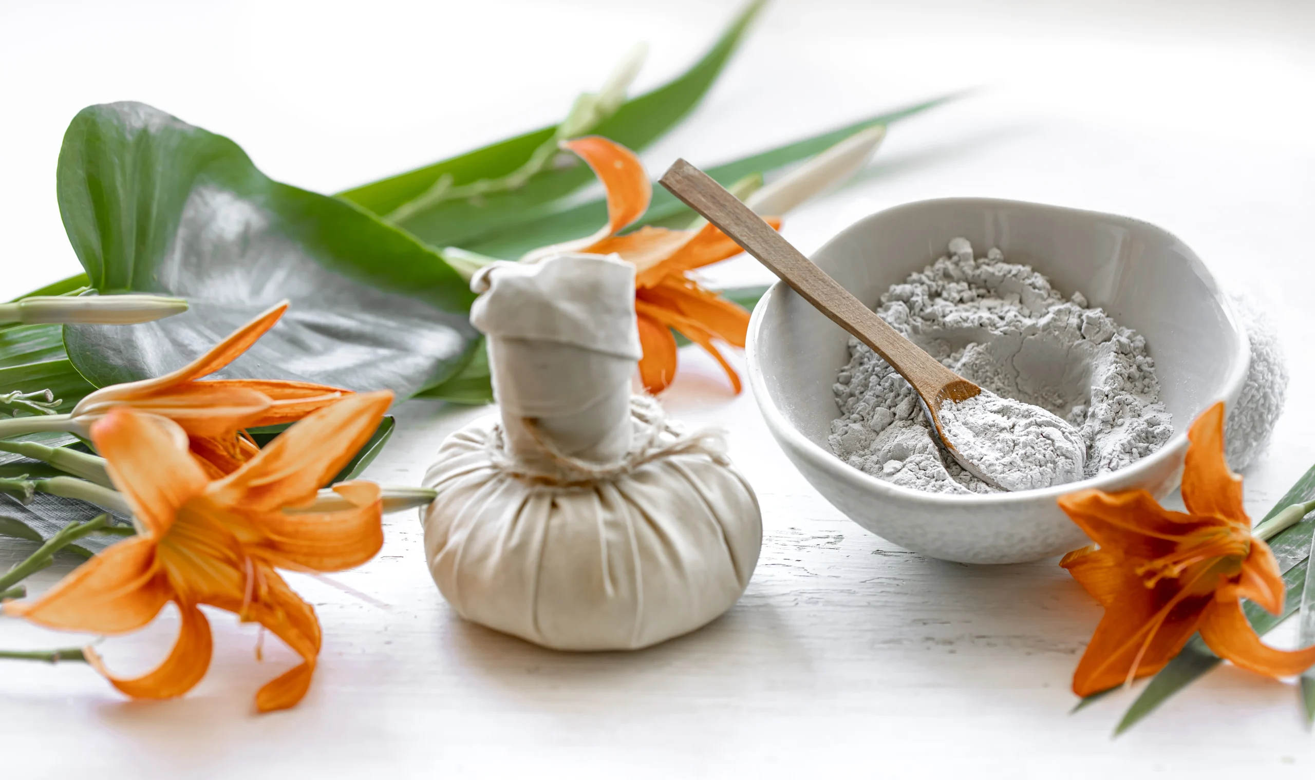 This image features a natural wellness setting centered on a traditional Kizhi (herbal poultice) bundle, a key tool used in Ayurvedic therapies for pain relief and inflammation. Beside the white fabric poultice, there is a ceramic bowl containing a fine grey herbal powder with a small wooden spoon, used for preparing therapeutic treatments. The arrangement is accented by vibrant orange lilies and lush green tropical leaves against a bright, clean white background, evoking a sense of holistic healing and professional care.