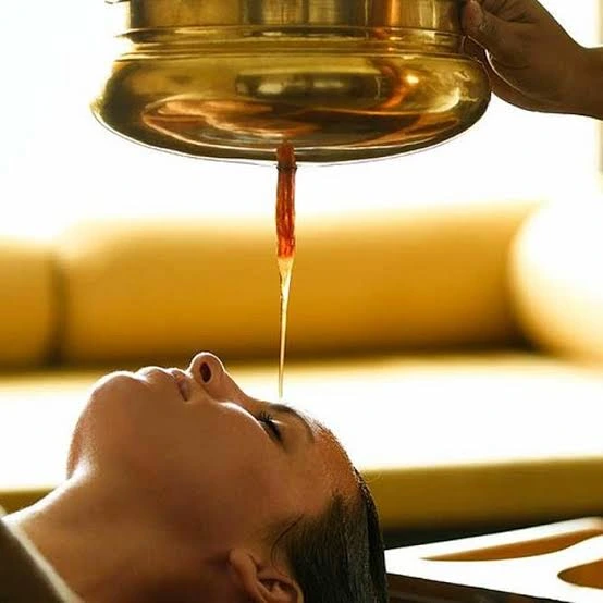 Woman receiving an Ayurvedic summer treatment with warm herbal oil gently poured onto her forehead from a brass vessel, promoting relaxation, cooling, and holistic wellness.