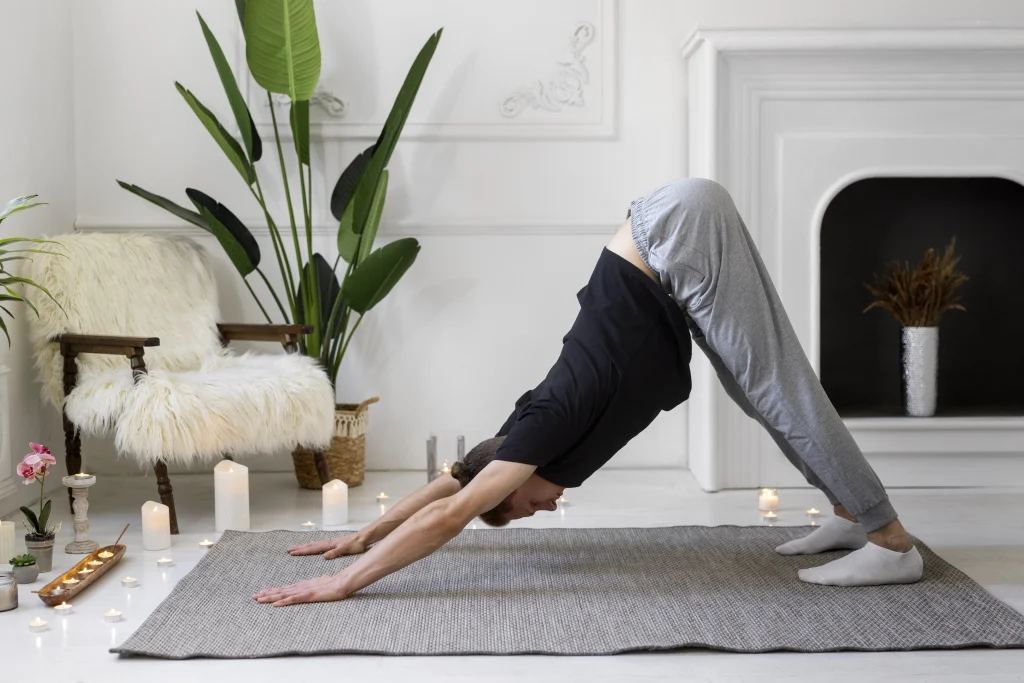Here are three alt-text options for the file full-shot-man-doing-yoga-mat.jpg: Short: Man practicing a yoga pose on a mat. Descriptive: A man in a black shirt and grey pants performing a downward-facing dog yoga pose on a mat. Contextual: A man doing yoga in a bright, minimalist room filled with plants and candles.