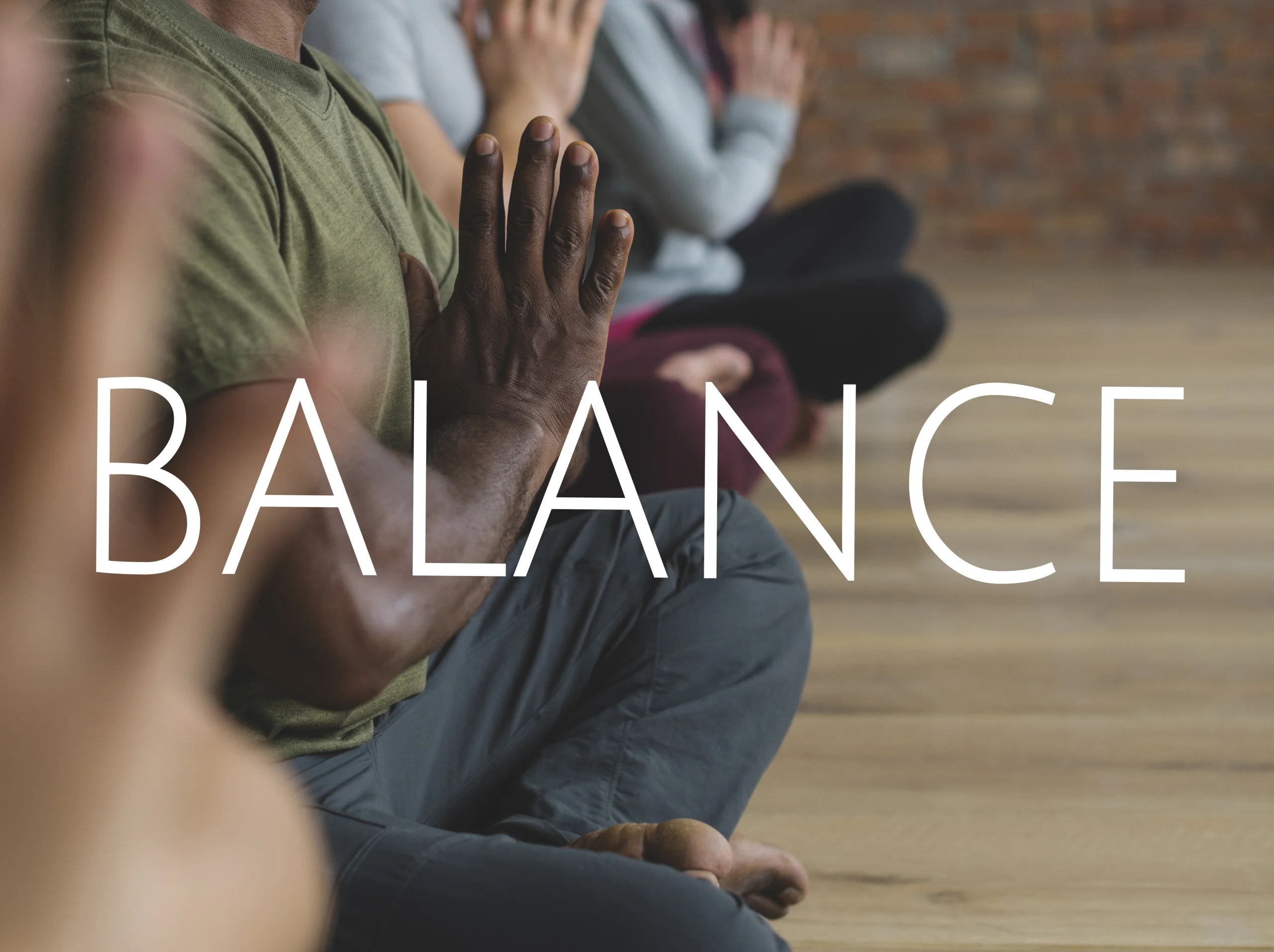 Integrated Wellness-A yoga class scene with people seated in meditation pose indoors, hands pressed together in prayer position. In the foreground, a participant with dark skin and green shirt is in focus while others are softly blurred in the background, and the word “BALANCE” appears in large white letters across the center of the image.